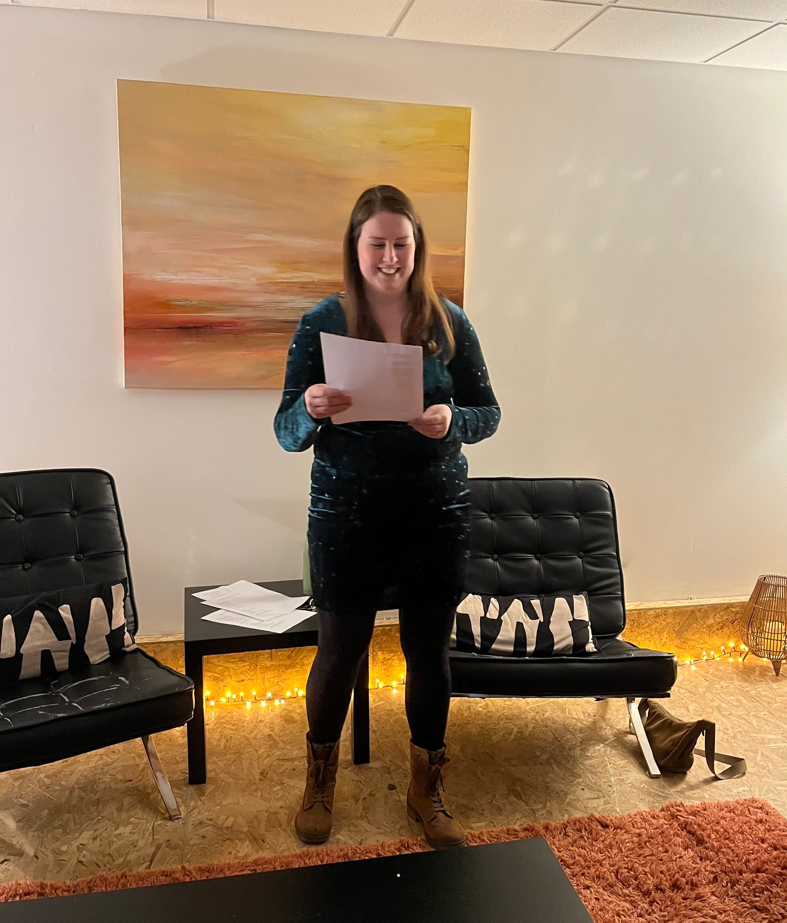 Smiling woman in a green dress reads from a paper between two black leather chairs.