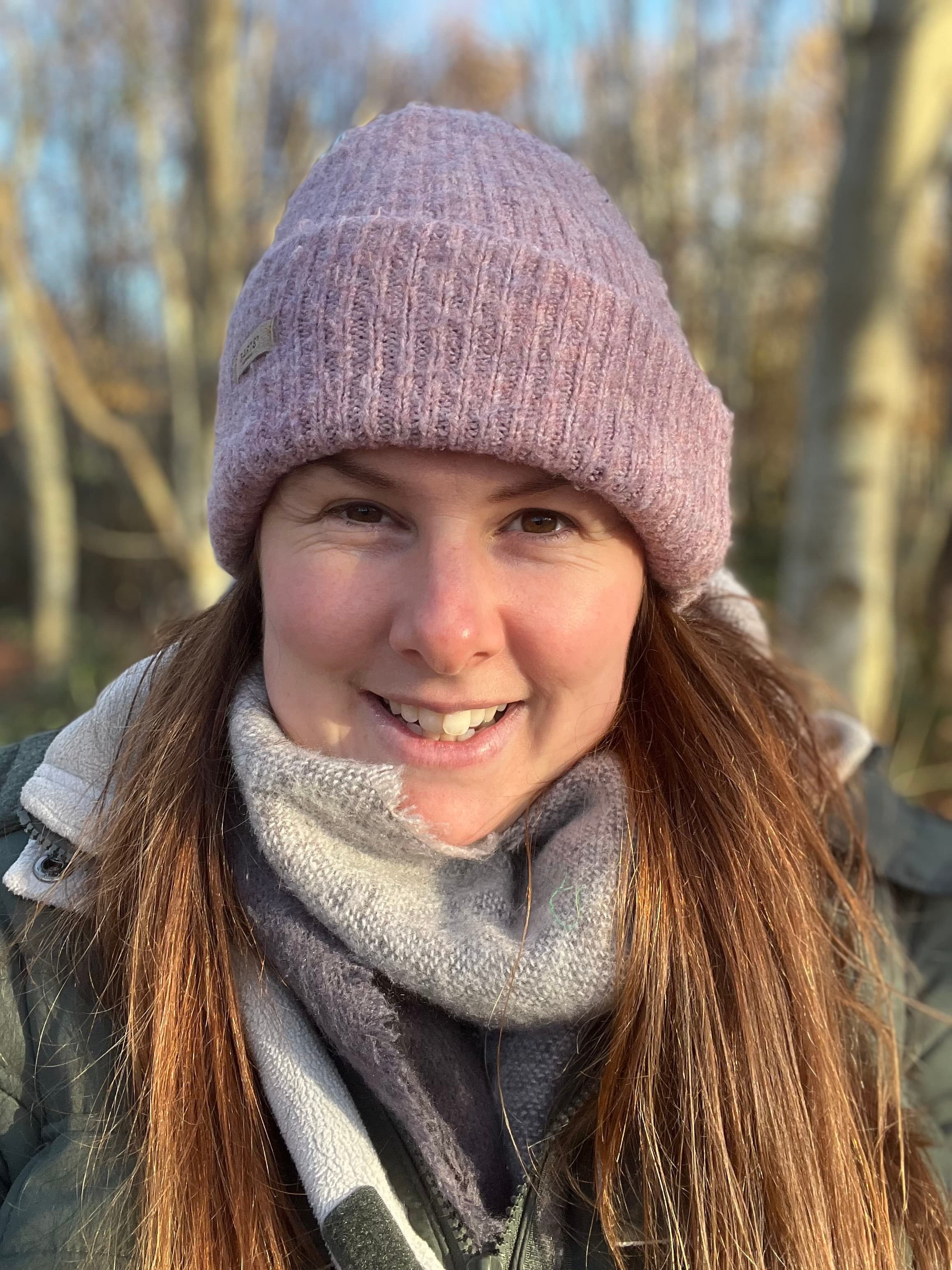Smiling woman wearing a purple knit beanie and grey scarf in a blurred forest.